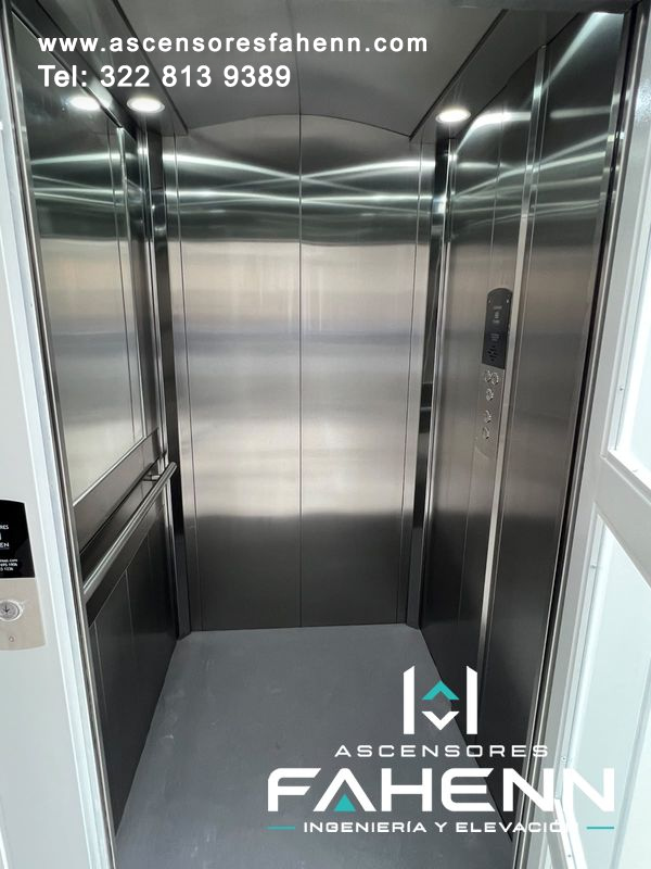 Cabina de ascensor en acero inoxidable con espejo lateral, pasamanos horizontal, botonera, puertas centrales y piso gris.