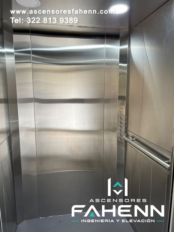 Ascensor residencial con cabina de acero, puertas centrales cerradas, pasamanos doble, botonera lateral y piso gris.