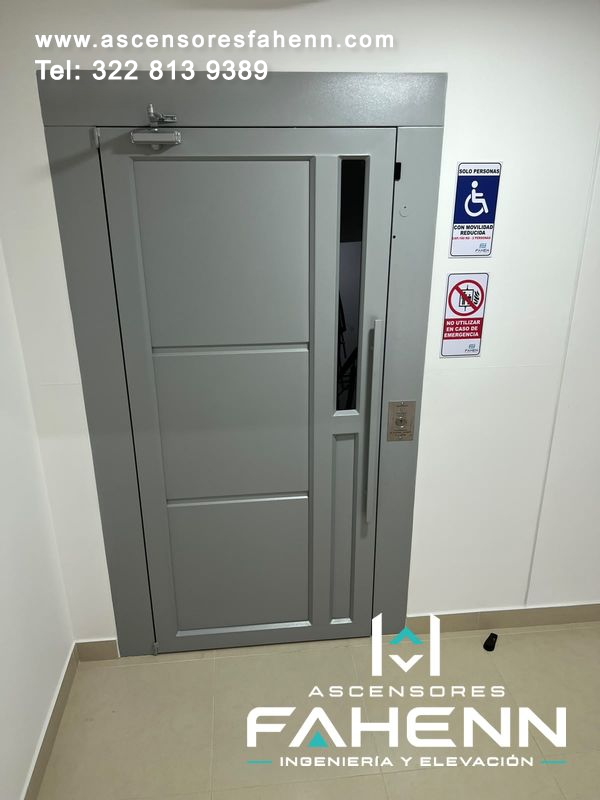 Ascensor para personas con movilidad reducida con puerta gris, visor vertical, tirador largo y señales de seguridad.