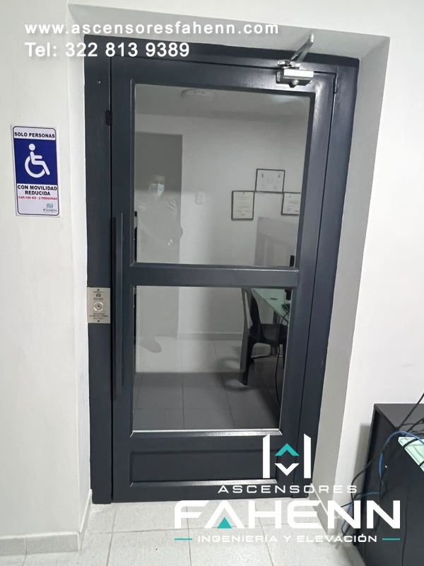 Plataforma elevadora accesible con puerta negra de vidrio, manija vertical, cierrapuertas y señal de movilidad reducida.