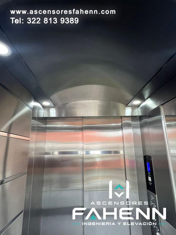 Cabina de ascensor en acero con puertas automáticas, techo curvo, luces circulares y botonera digital iluminada.