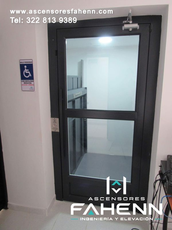 Ascensor accesible con puerta negra, dos paneles de vidrio, tirador vertical, cierr? purtas y señal para movilidad reducida.