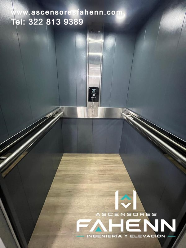 Cabina de ascensor moderno con paredes grises, pasamanos dobles, panel de control central y piso tipo madera.