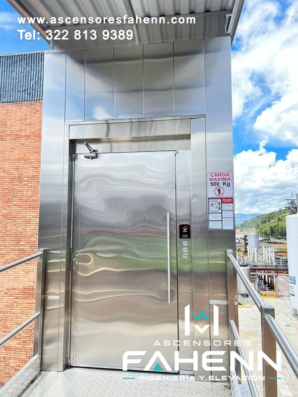 Ascensor de carga de 500 kg con puerta de acero, tirador vertical, cierrapuertas, botonera y señal informativa.