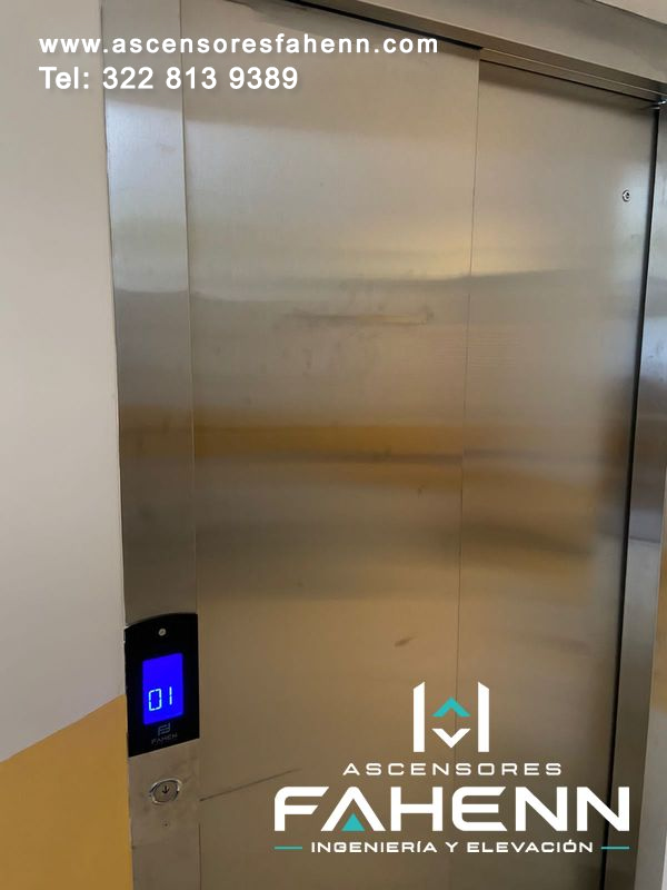 Puertas automáticas de ascensor en acero inoxidable con indicador digital azul del primer piso y pulsador exterior.