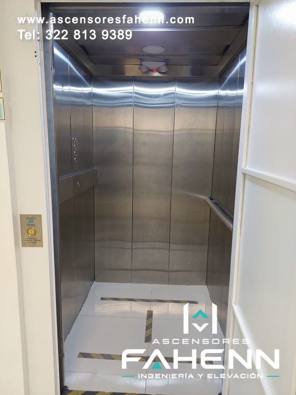 Cabina de ascensor moderno con paneles de acero, pasamanos lateral, botonera y piso claro con franjas de seguridad.