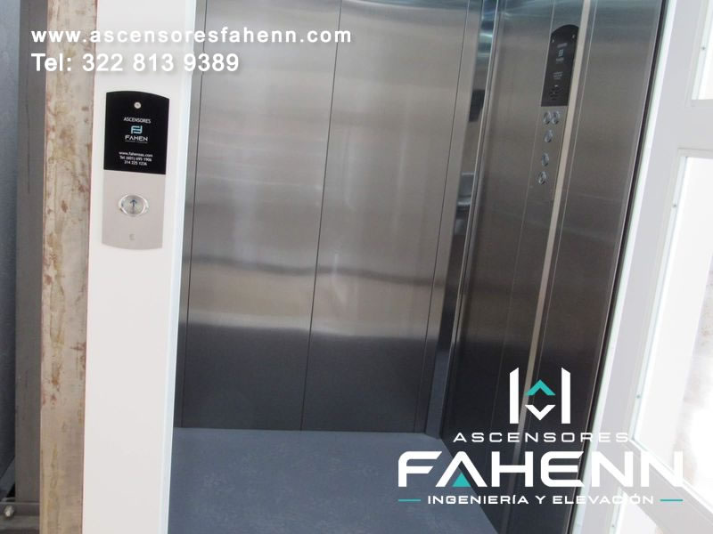 Ascensor para casa con cabina de acero, puertas centrales, botonera lateral moderna, pantalla y piso gris.
