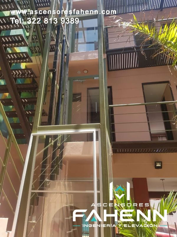 Ascensor exterior de vidrio instalado junto a corredores residenciales, con estructura vertical resistente que conecta pisos y mantiene luminosidad en el patio.