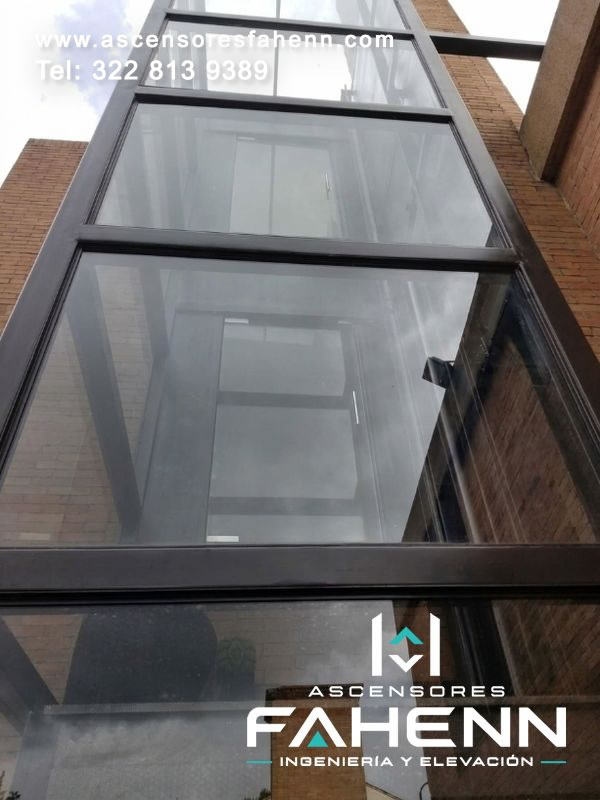 Ascensor panorámico instalado entre edificios con torre acristalada y perfiles oscuros, diseñado para conectar niveles y aprovechar un espacio vertical disponible.